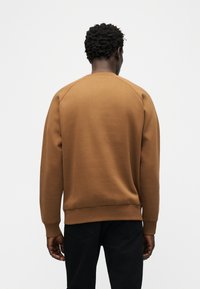 Άνδρας με κοντά σγουρά μαλλιά, φοράει καφέ φούτερ και μαύρο παντελόνι, στέκεται με την πλάτη προς τα εμπρός μπροστά σε απλό λευκό φόντο.