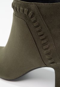 Gros plan d'une bottine en daim vert olive avec un détail de couture décorative en daim près du haut et un talon effilé.