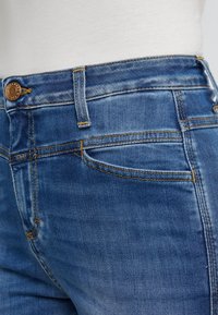 Jeans i medelblå tvätt, med en framficka, synlig sömnad och en metallknapp i midjan.