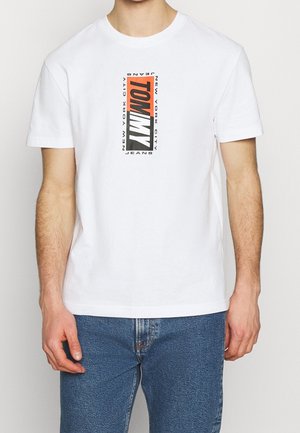 Mężczyzna noszący biały t-shirt z pionowym logo "Tommy New York City Jeans" w czerwonym, czarnym i białym kolorze, w zestawieniu z niebieskimi dżinsami.