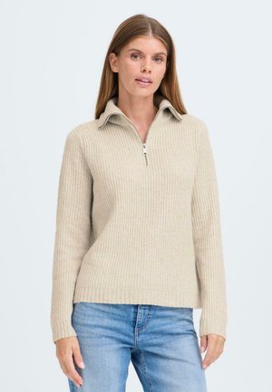 Pull en beige côtelé avec col zippé et manches longues, associé à un jean bleu. Le matériau semble doux et texturé.