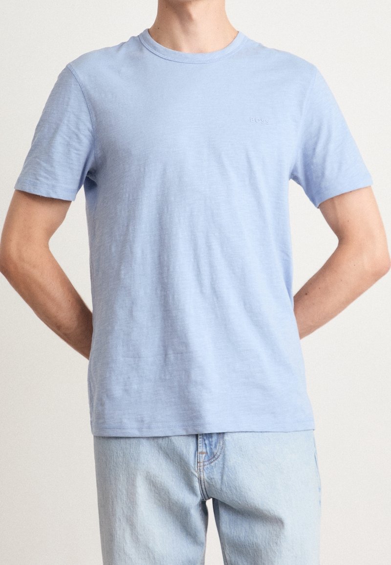 Homme portant un t-shirt bleu clair à manches courtes avec un logo discret sur la poitrine et un jean bleu clair, debout avec les mains derrière le dos.