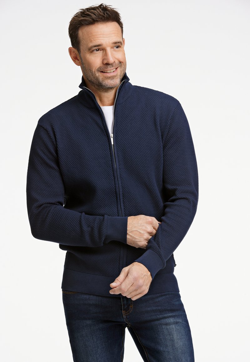 Navy zip-up jacka med en texturerad mönster, ribbade ärmmuddar och midjeband. Bärs över en vit skjorta, har en hög krage.