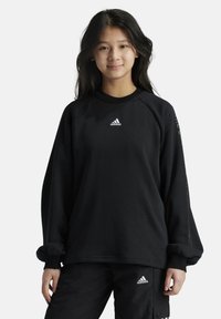 Sudadera negra de manga larga, con cuello redondo acanalado y logotipo blanco en el pecho. Presenta puños fruncidos y un corte relajado. Combinada con pantalones negros.