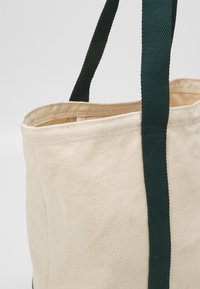 Sac fourre-tout en coton de couleur naturelle avec un tissu texturé, doté de poignées tissées vert foncé et d'un haut ouvert pour un accès facile.