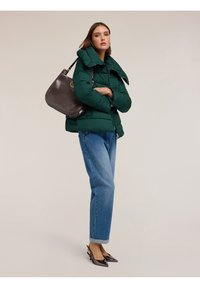 Zelená péřová bunda s vysokým límcem, v kombinaci se světle modrými džíny. Celý outfit doplňuje tmavě hnědá kožená kabelka a lesklé černé boty.