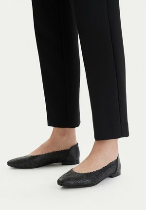 Stopy w czarnych balerinach z kwiatowym wzorem, zestawione z czarnymi spodniami o prostych nogawkach zakończonymi na wysokości kostek, na jednolitym tle.