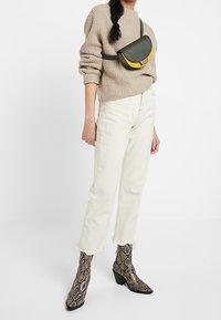 Kobieta w beżowym swetrze z dzianiny, kremowych krótkich jeansach, botkach w wę�żowy wzór i małej torebce na ramię z zieloną i żółtą klapką.