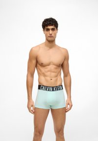 Jauns vīrietis ar īsām, lokainām matu frizūrām, tērpts gaiši zilos Calvin Klein bokseršortos, stāv uz balta fona.