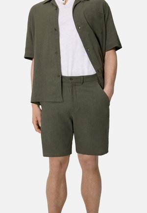 Hombre con camisa de manga corta color verde oliva abotonada sobre camiseta blanca, combinada con pantalones cortos a juego en verde oliva, con las manos en los bolsillos.