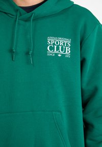 Green hoodie with a soft texture, featuring a front pocket, adjustable drawstrings, and white "ADIDAS ORIGINALS SPORTS CLUB" logo.