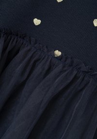 Abito blu navy con un top a costine decorato con motivi a cuore dorati e una gonna in tulle a strati con una cucitura arricciata. Texture morbida e design strutturato.