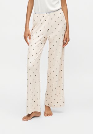 Vrouw die crèmekleurige pyjamabroek met brede pijpen en kleine donkere bloemenprint draagt, blootsvoets staat op een witte vloer, getoond van de taille naar beneden.