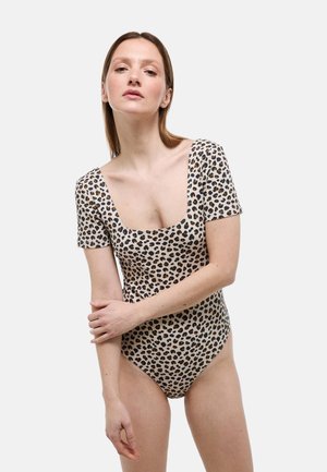 Femme aux cheveux bruns lisses portant un body à manches courtes et encolure carrée imprimé léopard, debout devant un fond blanc.