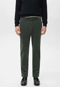 Mężczyzna ubrany w ciemnozielone sztruksowe spodnie, czarne skórzane buty i czarny sweter, stojący na tle jednolitej białej ściany.