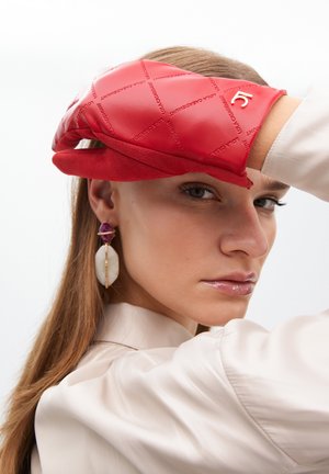 Guante de cuero rojo con diseño acolchado, que presenta un pequeño logotipo dorado, combinado con una camisa de seda beige y pendientes llamativos.