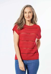 Rode gebreide top met korte mouwen, met horizontale uitsparingspatronen. Gecombineerd met blauwe jeans, die een gestructureerde afwerking hebben.