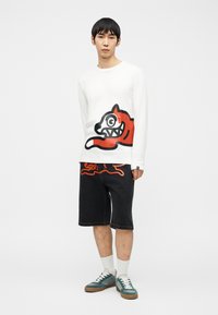 Νέος άνδρας που φοράει λευκό πουκάμισο μακρυμάνικο και μαύρο σορτς, και τα δύο με ίδιο κόκκινο σχέδιο καρτούν σκύλου, πράσινα αθλητικά παπούτσια και λευκές κάλτσες.