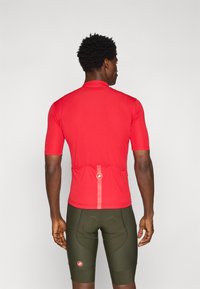 Maillot de cyclisme rouge avec un col court et des manches courtes, doté d'une poche arrière et d'accents contrastés. Porté avec un short vert olive.