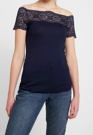 Vrouw draagt een marineblauwe off-shoulder top met kanten details op de mouwen en halslijn, gecombineerd met een blauwe spijkerbroek tegen een effen achtergrond.