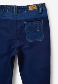 Pantalones de mezclilla azul marino con una cinturilla azul contrastante, que presentan costuras doradas y un bolsillo trasero con detalles decorativos en la costura.