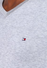 Hellgrauer V-Ausschnitt-Pullover aus einem weichen, strukturierten Stoff; mit einem kleinen, gestickten Rot-, Weiß- und Blau-Logo auf der Brust.