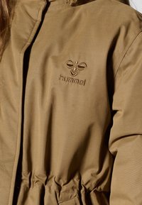 Veste isolante marron à taille cintrée, avec un logo brodé dans le coin supérieur gauche et une texture lisse.