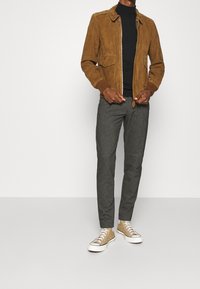 Bruine suède jas met een rits aan de voorkant, een zwarte coltrui eronder, grijze geruite broek en beige sneakers met witte zolen.