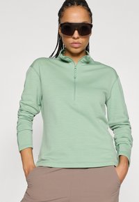 Ανοιχτό πράσινο μακρυμάνικο πούλοβερ με ψηλό γιακά και μισό φερμουάρ, κατασκευασμένο από υφασμάτινη υφή. Συνδυάζεται με καφέ παντελόνι και σκούρα γυαλιά ηλίου.