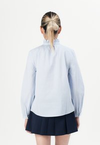 Kvinne med lyst hår i hestehale iført en blå og hvit stripete langermet bluse og et marineblått plissert skjørt, sett fra bak.