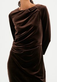 Långärmad klänning i mörkbrunt sammetstyg, med en draperad framdel, slät yta och figurvänlig silhuett med sidofickor.