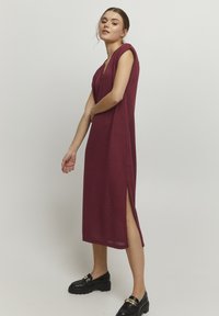 Robe bourgogne, longueur genou, avec un décolleté en V et de courtes manches. Présente une fente sur le côté et un tissu texturé. Associée à des mocassins noirs à semelles épaisses.