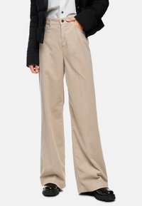 Beige wide-leg corduroy pants with a button front, front pockets, and a textured finish. Paired with a black puffer jacket and shoes.