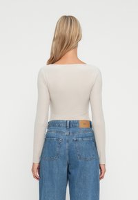 Eine blonde Person mit langen Haaren trägt ein enges, beiges Langarmoberteil, das in hochgeschnittene blaue Jeans gesteckt ist, zu sehen von hinten vor einem weißen Hintergrund.