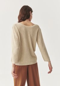 Beige kötött pulóver széles nyakkivágással, hosszú ujjakkal és bordázott textúrával. A háta enyhén oversize, és a derék alá ér.