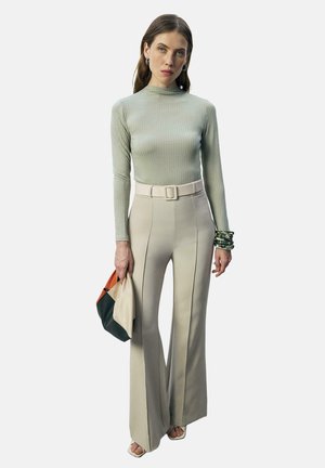 Femeie purtând un pulover verde deschis, mulat, cu dungi, și pantaloni evazați bej cu talie înaltă, ținând un plic multicolor și purtând brățări.