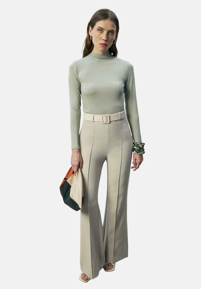 Γυναίκα που φοράει εφαρμοστό ανοιχτοπράσινο πλεκτό πουλόβερ με ρίγες και ψηλόμεσα μπεζ παντελόνια καμπάνα, κρατώντας πολύχρωμο clutch και φορώντας βραχιόλια.