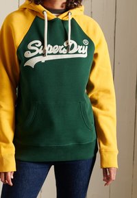 Felpa verde e gialla con cappuccio, caratterizzata dal logo "SuperDry" in bianco, tasca frontale e polsini a costine. Tessuto morbido.