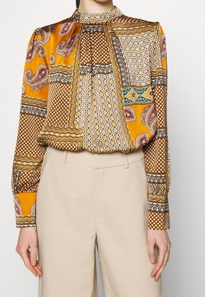 Kvinde iført en bluse med høj hals og lange ærmer med flerfarvet patchworkmønster, kombineret med beige bukser med høj talje.