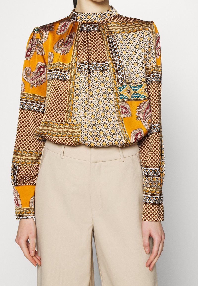 Femme portant un chemisier à col montant et manches longues avec un motif patchwork multicolore, associé à un pantalon beige taille haute.