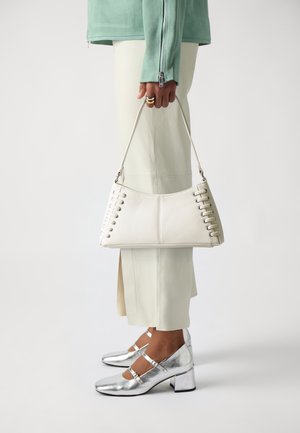 Hvid læderhåndtaske med snøre detaljer på siden, båret af en person. Sølvfarvede sko med blokhæle fuldender outfittet.