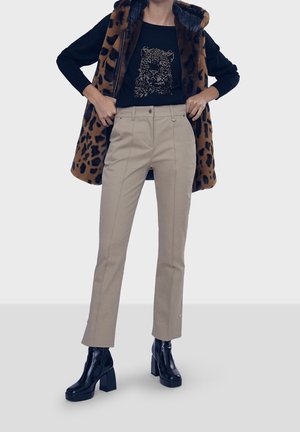 Kvinde iført beige skræddersyede bukser, sorte støvler, sort skjorte med leopardansigtsprint samt leopardmønstret ærmeløs pelsjakke.