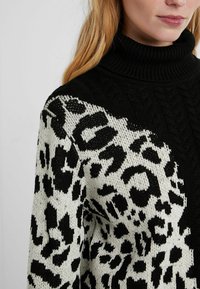Pull à col roulé noir avec un tricot texturé, présentant un imprimé léopard en noir et blanc sur une manche et la zone de l'épaule.