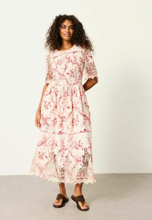 Woman with curly hair wearing a white and red floral midi dress with lace sleeves and hem, standing on a gray floor.