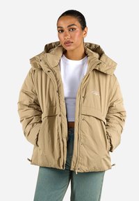 Beige isolierte Pufferjacke mit Kapuze, ausgestattet mit einem Reißverschluss, zwei Fronttaschen und einem dezenten Logo auf der Brust. Texturiert.
