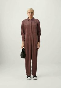 Bordo kordfløyels jumpsuit med knapper foran, lange ermer og to frontlommer. Brukt med en liten svart veske og svarte joggesko.