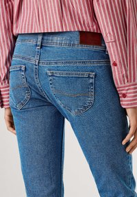 Blå denimjeans med midje i middels høyde, med to baklommer og kontrasterende sømmer. Brukt sammen med en rødt og hvitt stripete skjorte.