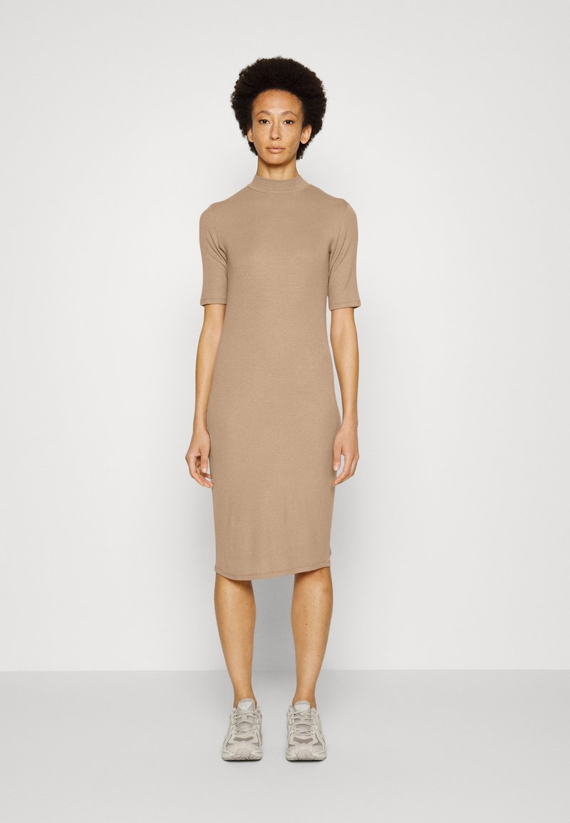 Vestido de malha, de comprimento midi, em bege claro com decote alto e mangas curtas. Usado com ténis cinza claro. Textura suave, design ajustado.