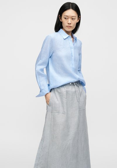 Femme portant une chemise bleu clair boutonnée, rentrée dans une jupe rayée taille haute avec de grandes poches avant, debout les mains dans les poches.