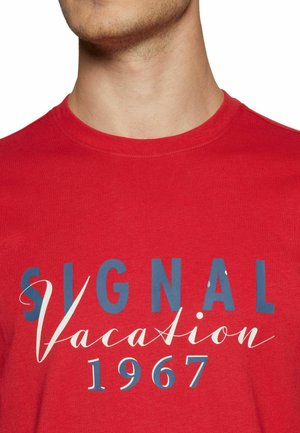 Mand iført en rød t-shirt med rund hals og teksten "SIGNAL Vacation 1967" trykt i blåt og hvidt på brystet.